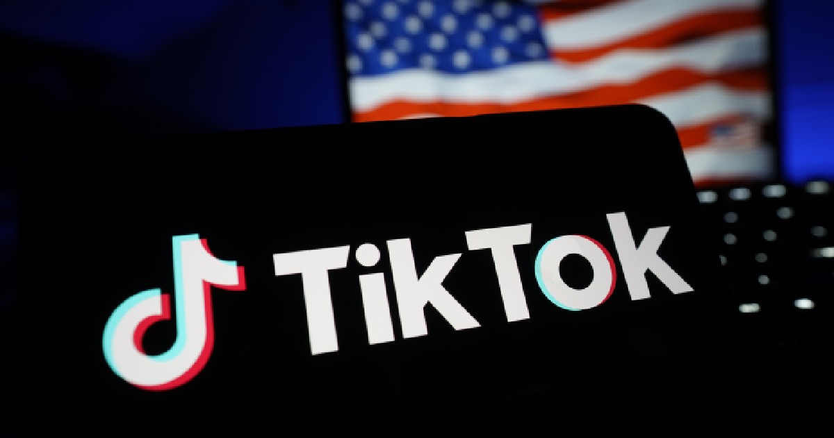 Imaginea surprinde logo-ul aplicației de social media TikTok afișat pe ecranul unui dispozitiv mobil, contrastând puternic cu fundalul întunecat. În fundalul nefocalizat, se distinge parțial un steag al Statelor Unite ale Americii pe ecranul unui alt dispozitiv.
