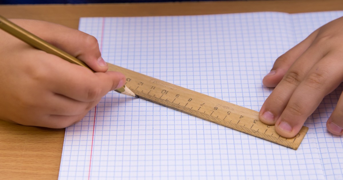 Un elev folosește o riglă din lemn pentru a trasa o linie pe o foaie de matematică cu pătrățele. Imaginea surprinde un moment obișnuit din timpul unei activități școlare, evidențiind utilizarea instrumentelor clasice de măsurare și desen.