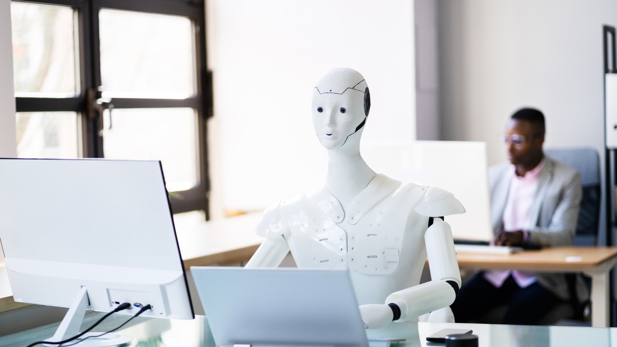 Imagine cu un robot alb stând la birou în fața unui laptop și a unui monitor. În spate se află un alt om care privește într-un monitor