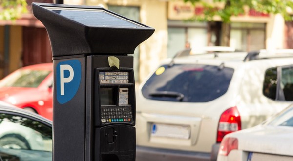 Prim-plan cu un aparat electronic de plată a parcării, alimentat cu panou solar, amplasat pe marginea unei străzi cu mașini parcate.