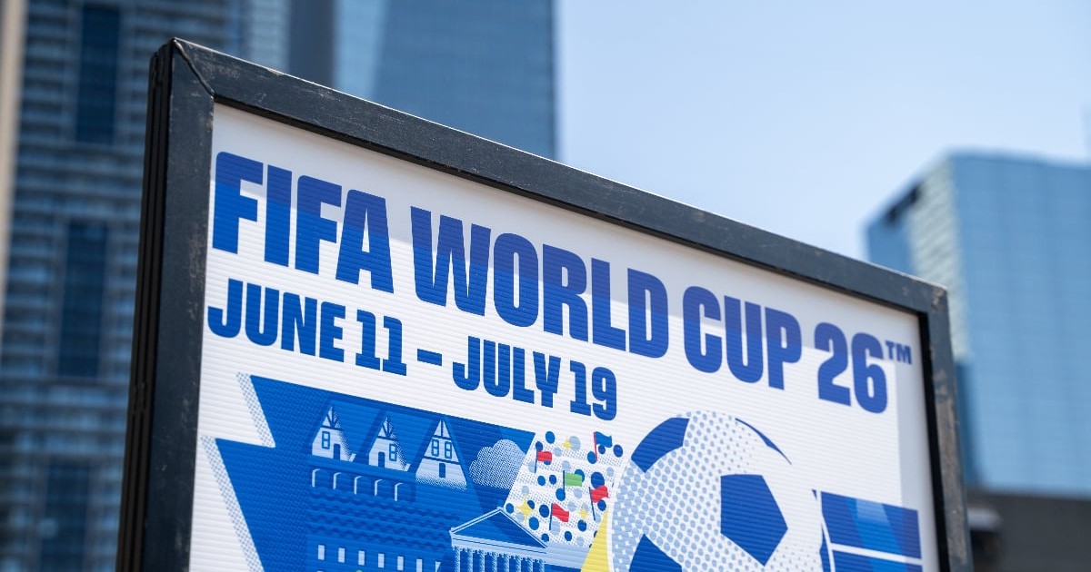 Imaginea prezintă un panou informativ înrămat, inscripționat cu textul "FIFA WORLD CUP" și datele "JUNE 11 – JULY 26", amplasat într-un cadru urban cu zgârie-nori moderni în fundal. Pe partea grafică a panoului se remarcă o minge de fotbal stilizată și o reprezentare grafică alb-albastră a unor clădiri și a unui peisaj.