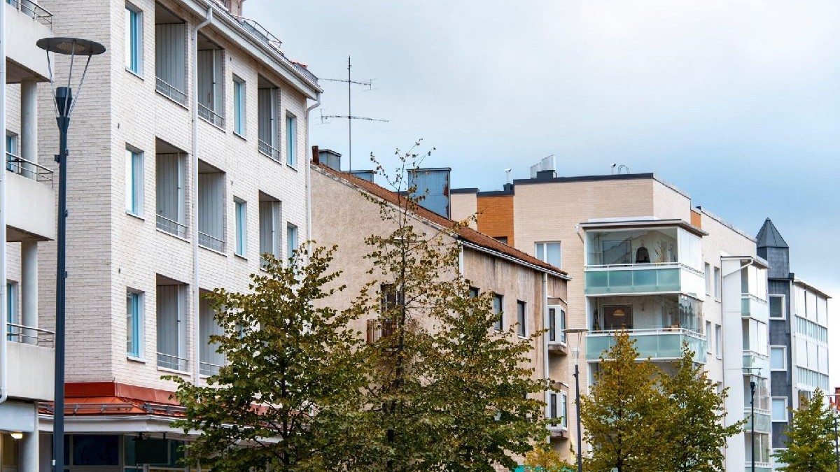 Fotografie cu o stradă urbană modernă, mărginită de clădiri de culoare bej și crem, cu balcoane și ferestre din sticlă. În prim-plan se află copaci verzi și un stâlp de iluminat