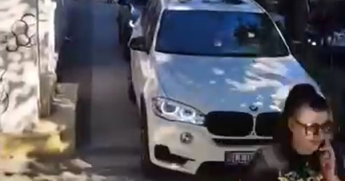 O femeie vorbește la telefon mergând pe trotuar, în spatele ei circulă o mașină albă