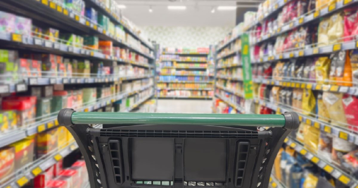 Prim-plan cu un cărucior de cumpărături, pe culoarul unui supermarket, cu cu raftru de o parte și de alta