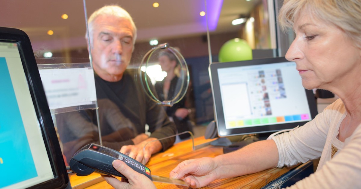 n interior în care o femeie cu părul scurt, blond, îmbrăcată într-o bluză crem, este surprinsă din profil în timp ce ține un terminal de plată negru și un card. Ea se află în fața unui panou despărțitor din sticlă prevăzut cu o deschidere circulară, în spatele căruia se află un bărbat cu părul grizonat, purtând o bluză neagră și privind în jos. Pe tejgheaua din lemn sunt vizibile două monitoare, unul în stânga jos și altul în spatele femeii, acesta din urmă afișând o interfață digitală cu elemente grafice colorate