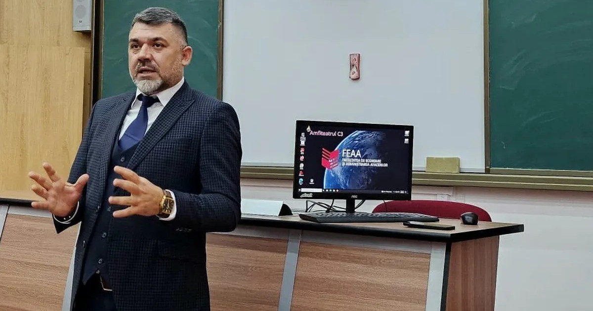 Costel Grojdea predă studenșilor fiind îmbrăcat într-un costum de culoare închisă, asortat cu o cravată iar în spatele său se află un ecran. Foto: Facebook- Costel Grojdea