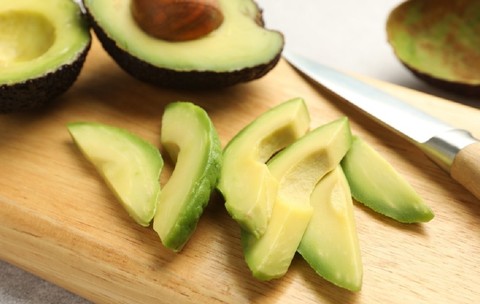 Avocado tăiat în jumătăți și felii așezate pe un tocător din lemn, alături de un cuțit de bucătărie cu mâner de lemn, pe un fundal deschis