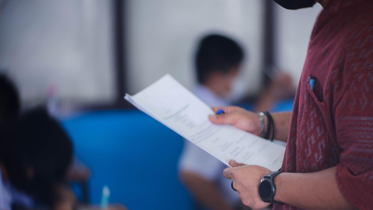 Imagine cu un tânăr care ține în mâini o foaie de examen. Poartă un tricou roșu închis și un ceas.