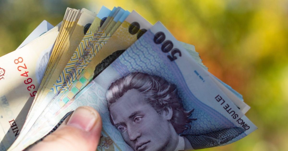 Bancnote de 500 și 50 de lei ținute într-o mână, iar pe fundal se pot observa, în blur, frunzele unui copac, de culoare verde și galbenă