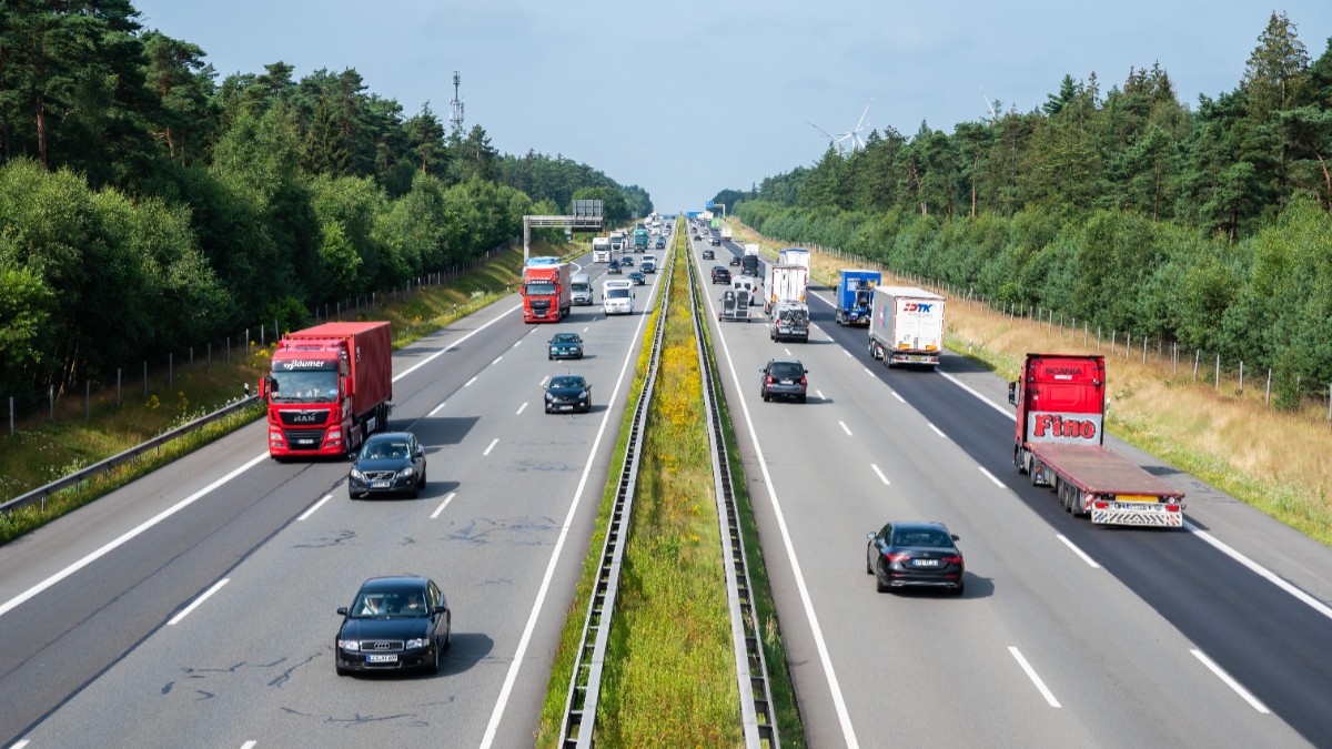 Imaginea prezintă o privire de sus asupra unei autostrăzi aglomerate, pe care circulă numeroase camioane și autoturisme în ambele sensuri, traversând o zonă împădurită.