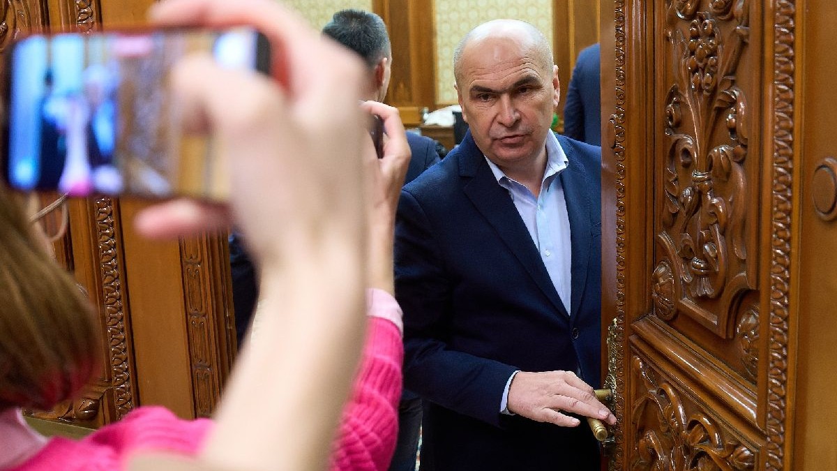 Fotografie cu premierul Ilie Bolojan, într-un costum bleumarin și cămașă albastră, lângă o ușă din lemn întredeschisă.
