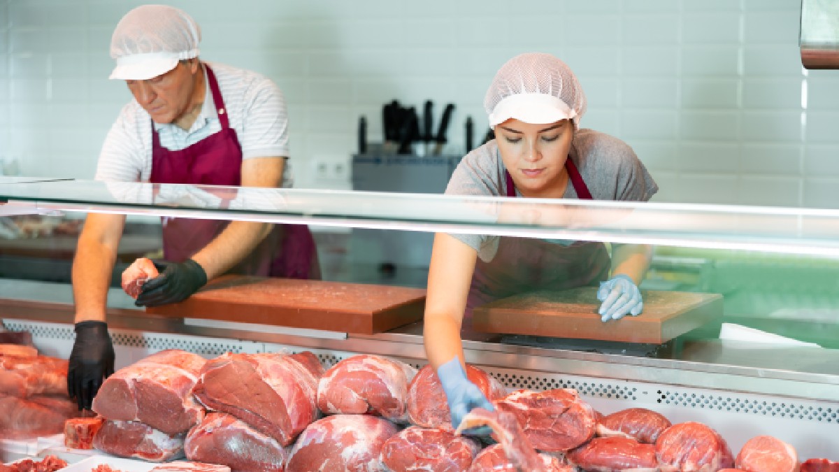 Imaginea surprinde doi măcelari purtând echipament de protecție sanitară în timp ce aranjează și pregătesc diverse sortimente de carne proaspătă în vitrina frigorifică a unui magazin de specialitate.