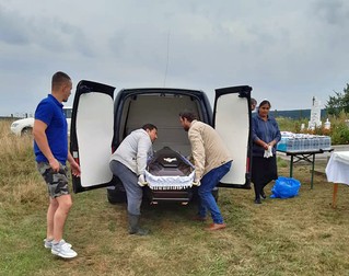 În albastru, angajatul firmei de pompe funebre, care nu poartă niciun costum de protecție și e în pantaloni scurți, privește un fiu al Anei Nagy și un prieten de familie coborând sicriul din mașină