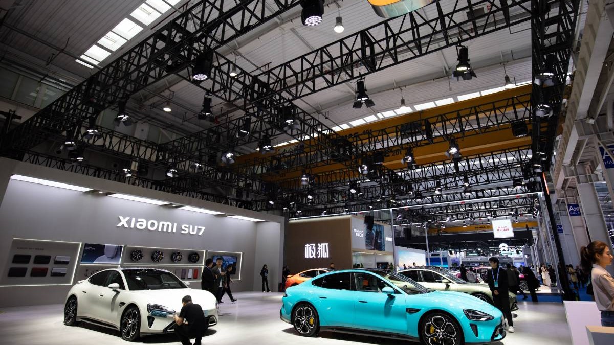 Showroom auto unde sunt expuse mai multe mașini electrice Xiaomi SU7. În prim plan sunt două modele electrice, unul alb și unul turcoaz. Printre mașini sunt cateva persoane care analizează părți ale fiecărei mașini