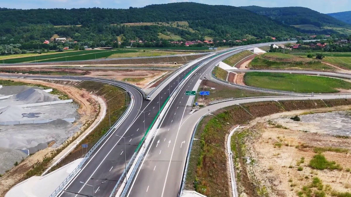 Imaginea oferă o vedere aeriană asupra unui nod rutier de autostradă, traversând un peisaj rural cu dealuri împădurite sub un cer senin.