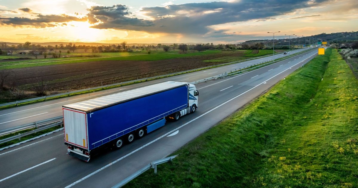 Un camion mare de transport merge pe o autostradă și trece printr-o zonă rurală, iar în stânga se vede sparele la apus