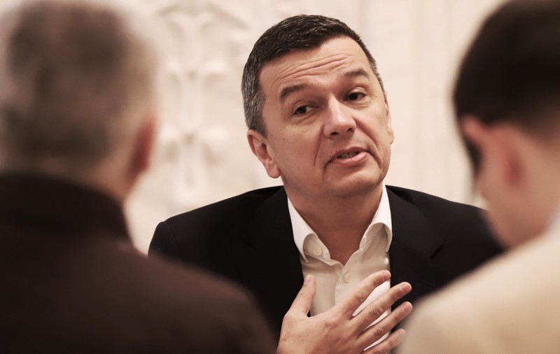 sorin grindeanu, cu mana pe piept și capul ușor într-o parte, purtând un costum negru cu cămașă albă, văzut printre umerii a alți doi bărbați îmbrăcați în costum