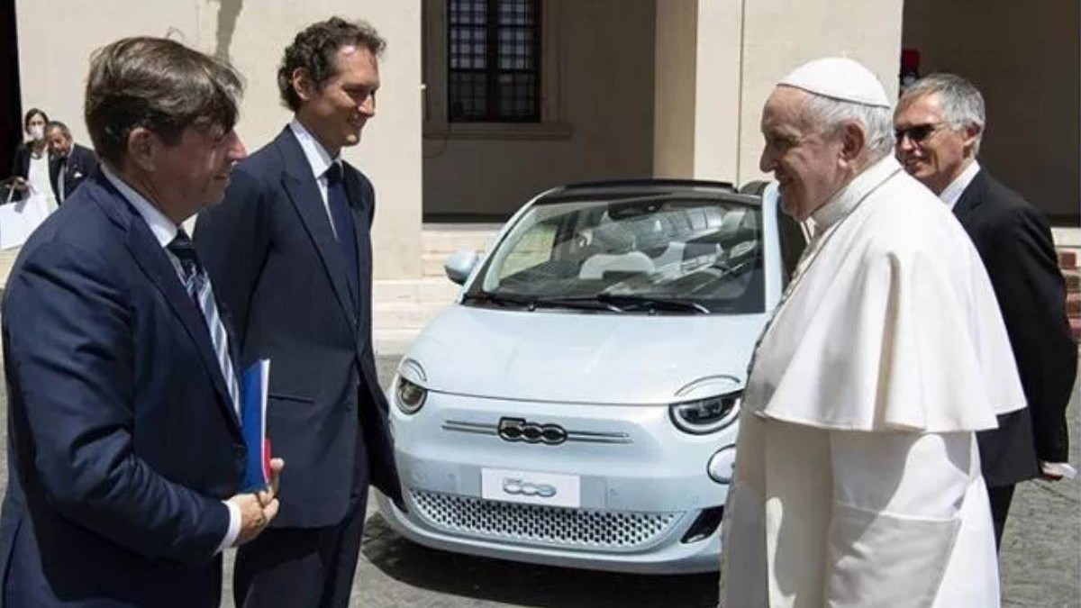 Papa Francisc îmbrăcat cu tradiționala vestimentație albă, în timpul unei discuții cu mai mulți oficiali, în fața unei mașini electrice Fiat 500 de culoare albă.