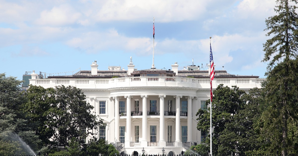 Imaginea prezintă fațada sudică a Casei Albe din Washington D.C., o structură neoclasică albă cu un portic semicircular, sub un cer parțial înnorat. Clădirea este încadrată de copaci verzi, iar în prim-plan este vizibil un gard metalic negru, cu steagul SUA fluturând în dreapta.