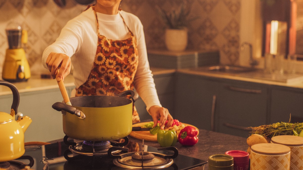 O femeie care face de mâncare la aragaz, în timp ce toacă legume și poartă un șorț cu flori
