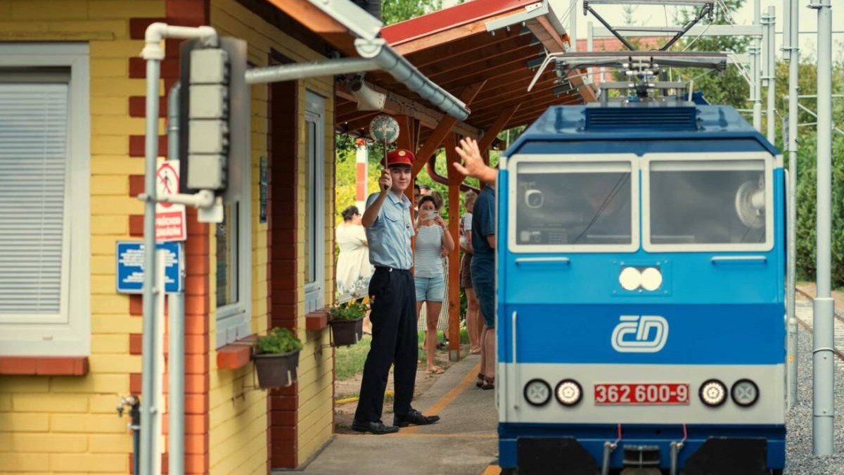 În imagine se vede unul dintre trenurile în miniatură ale rețelei Železnice 600, o locomotivă albastră oprită lângă un peron amenajat în grădină. În stânga, un bărbat îmbrăcat în uniformă feroviară și cu șapcă roșie face semn cu mâna, iar în spatele lui se află mai mulți vizitatori. În jurul trenului sunt vizibile clădiri de gară, stâlpi și instalații care imită infrastructura unei căi ferate reale.