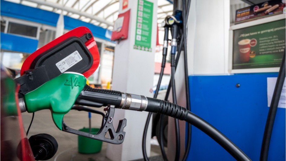 Imaginea prezintă un pistol verde de pompă de carburant introducător în rezervorul deschis al unei mașini roșii la o stație de alimentare.   
