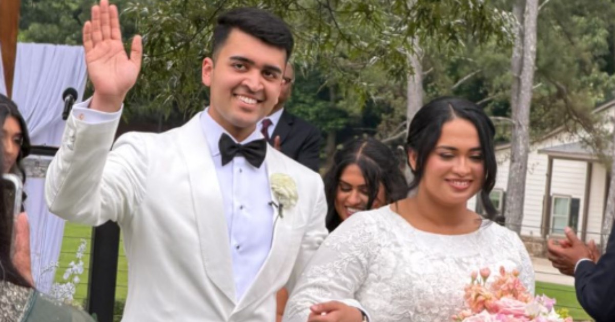 În imagine apare un cuplu de miri surprins în ziua nunții, în timpul unei ceremonii desfășurate în aer liber. Mirele salută invitații cu un zâmbet larg, în timp ce mireasa ține un buchet de flori și îl însoțește într-un moment plin de emoție și bucurie.
