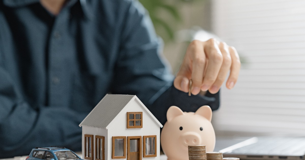 Imaginea prezintă o scenă simbolică legată de taxe și finanțe personale. În prim-plan se află o machetă de casă, o pușculiță în formă de porc și mai multe stack-uri de monede, sugerând economii sau costuri în creștere. Lângă acestea se vede o mașinuță miniaturală, reprezentând impozitarea auto.