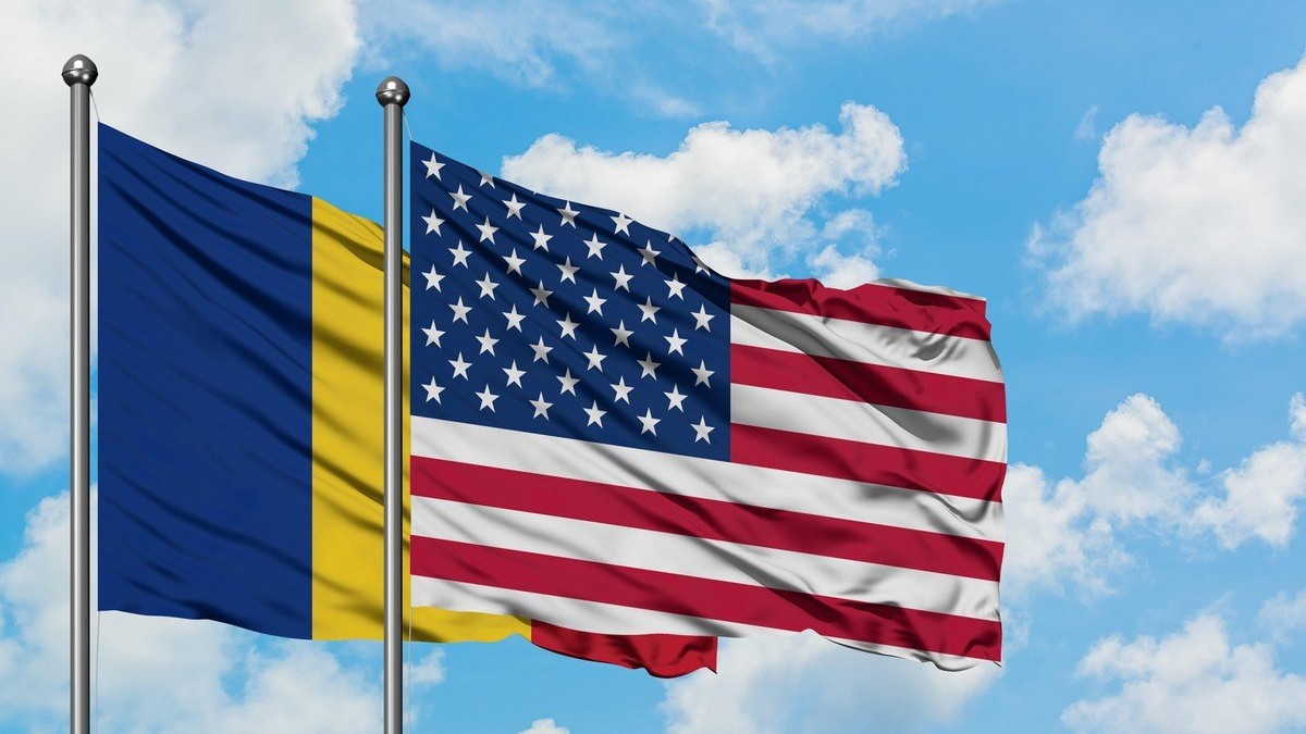 Steagurile României și Statelor Unite fluturând în vânt pe fondul unui cer albastru înnorat.