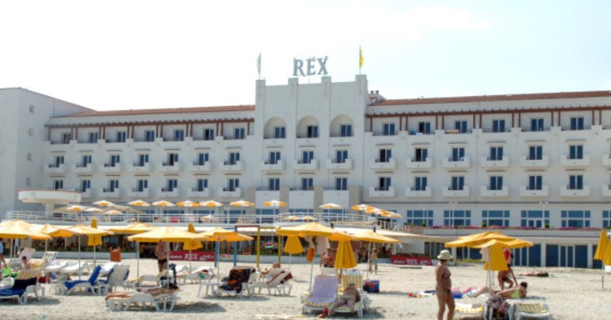 Imagine de pe plajă, cu oameni în prim plan și șezlonguri, iar pe fundal clădirea în care funcționează hotelul Rex din Mamaia