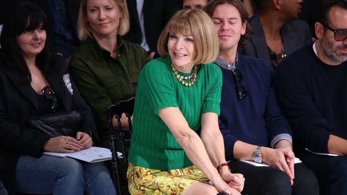Anna Wintour, așezată în primul rând la un eveniment de modă în Paris, septembrie 2018. Este îmbrăcată un pulover verde cu mâneci scurte, din material tricotat, și o fustă scurtă cu imprimeu floral galben. Stă cu picioarele încrucișate și mâinile împreunate în poală. În jurul ei sunt și alte persoane, care sunt îmbrăcate în culori reci