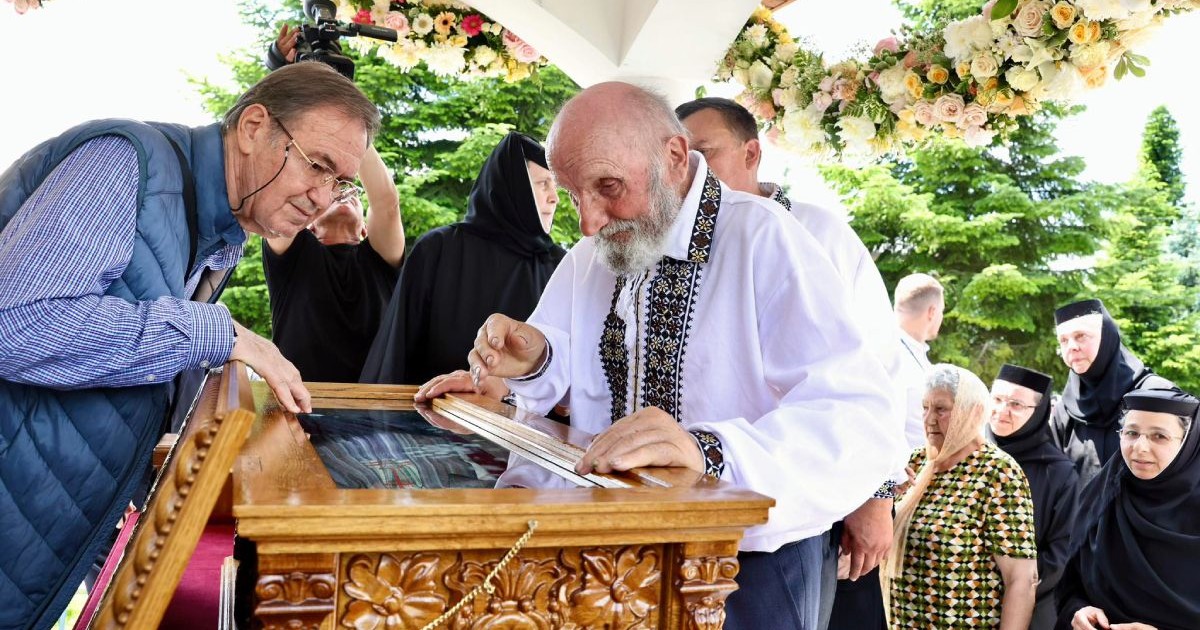 Vasile Lazăr, tatăl Sfintei Elisabeta de la Pasărea, îmbrăcat cu o ie românească, lângă racla cu moaștele fiicei sale, Sfânta Elisabeta de la Pasărea și alți oameni pe fundal