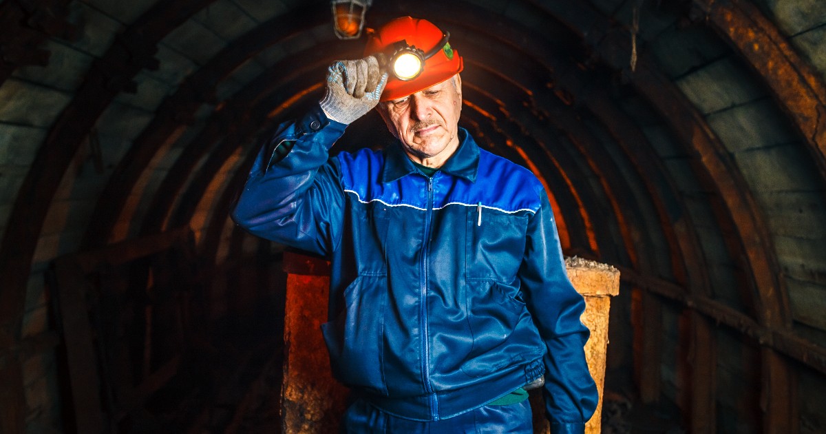 Un miner, echipat cu cască și lampă frontală, stă într-un tunel subteran slab luminat. Îmbrăcat în salopetă de lucru, acesta pare concentrat și obosit, sugerând condițiile dificile din mină.