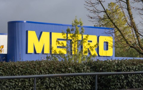Sigla METRO, cu litere galbene strălucitoare pe clădirea albastră în spatele verdeții luxuriante