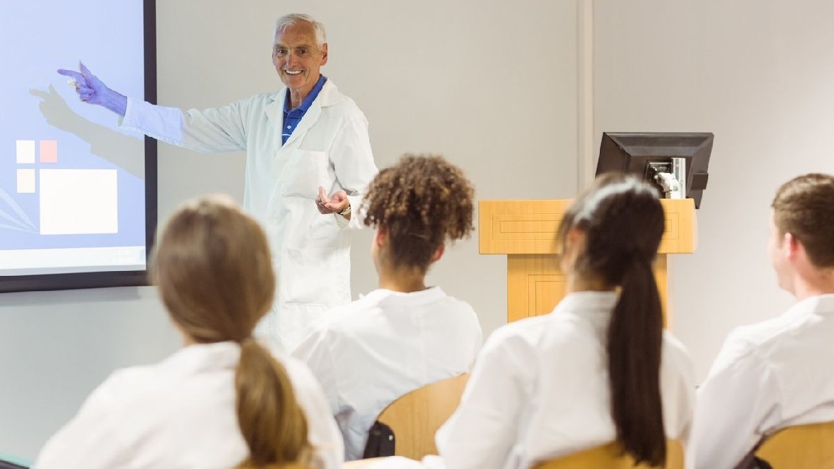 Un profesor arata ceva la tabla, patru studenti care stau cu fata spre tabla