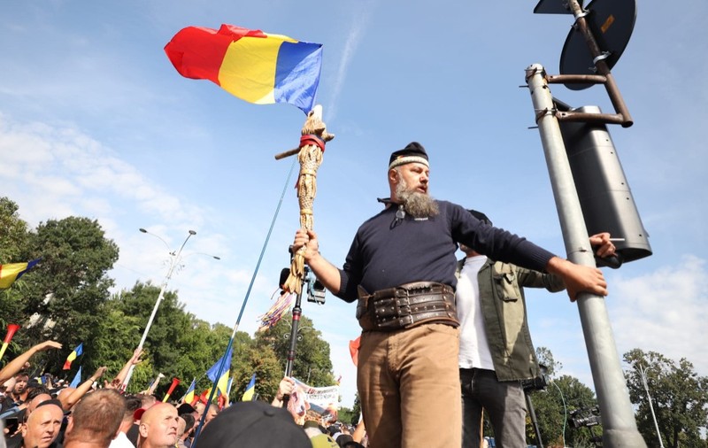Un bărbat cu barbă și brâu din piele, purtând un toiag decorat și un steag al României, urcat la înălțime deasupra unei mulțimi de protestatari.