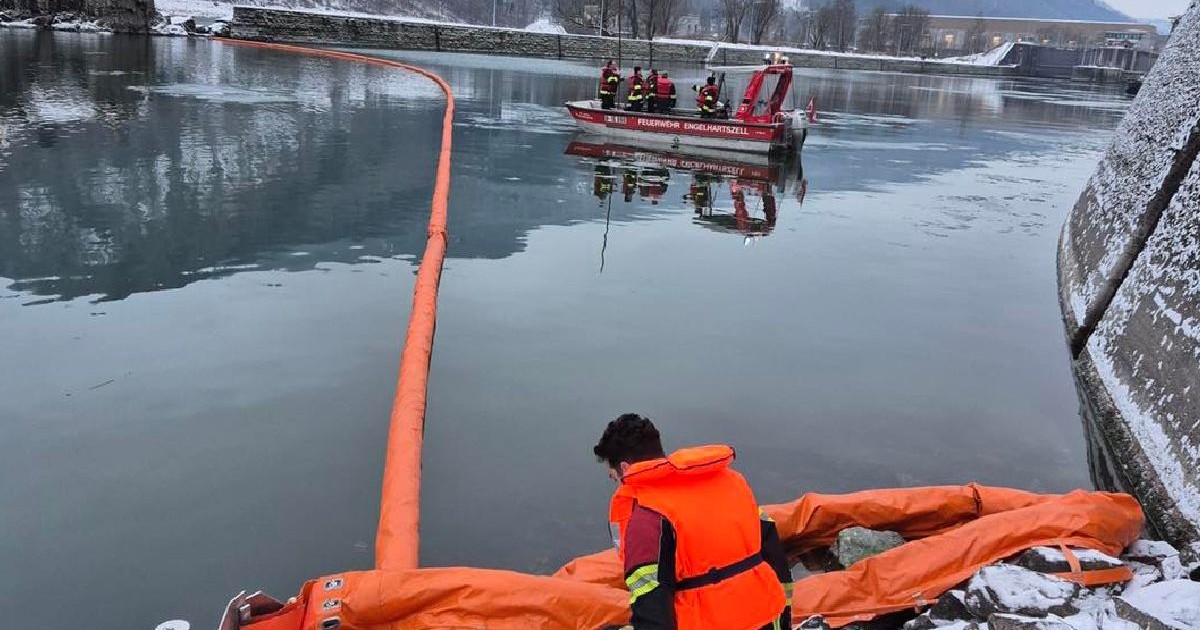 O echipă de salvare îmbrăcată în echipament portocaliu lucrează pentru a asigura o barcă gonflabilă de-a lungul malului înghețat al râului, cu o barcă de salvare roșie în apropiere. Pietre și copacii acoperiți de zăpadă înconjoară scena.