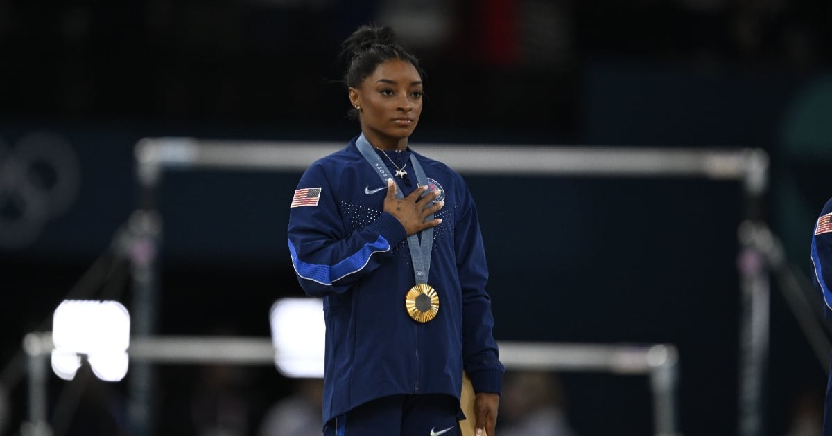 Simone Biles cu o medalie de aur la gât. Aceasta ține mâna dreapta la piept.