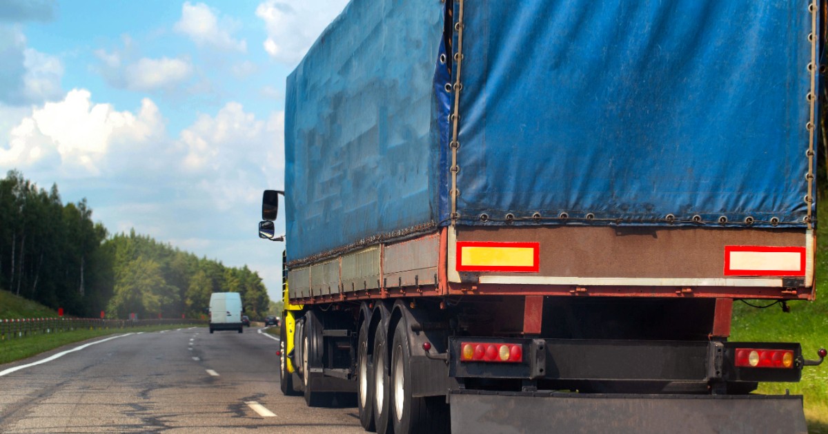 Prim-plan cu un camion care are prelată albastră și circulă pe partea dreapta a drumului, cu o dubă albă în față și copaci pe partea stângă a șoselei cu două benzi