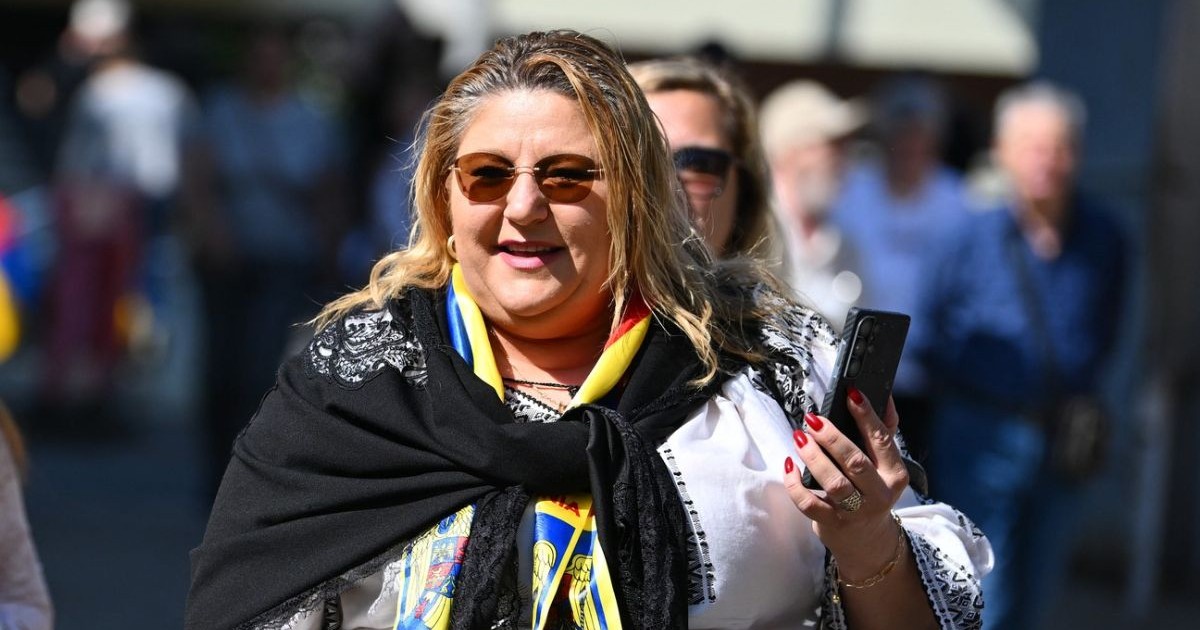 Diana Sosoacă intr-o ipostază pe stradă, rade, cu telefonul în mana stanga, care este ridicata, poartă o bluză alba cu model traditional romanesc si o eșarfă neagră legata la gat, oameni in spatele ei