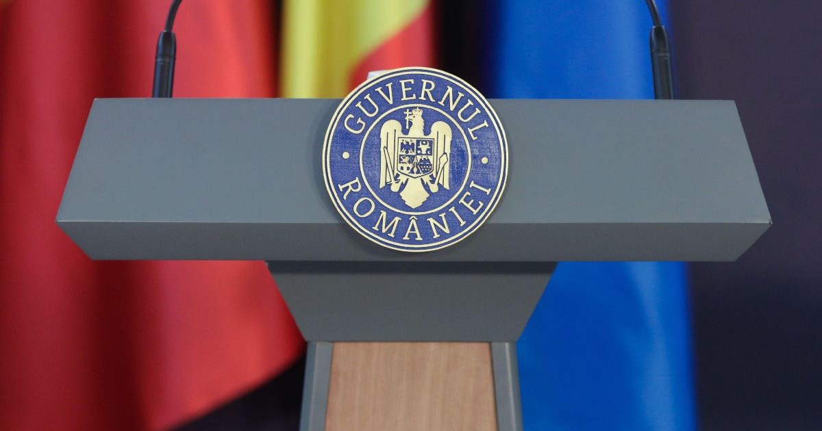 Imaginea prezintă în prim-plan o tribună oficială de culoare gri pe care este aplicată stema cu inscripția Guvernul României, având în fundal steaguri oficiale parțial vizibile.