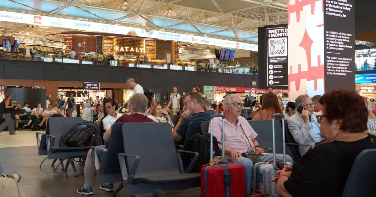 Terminalul aglomerat al aeroportului Fiumicino, Italia - 19 iunie 2026, plin de călători așezați pe scaune, bagaje în apropiere și magazine vizibile în fundal