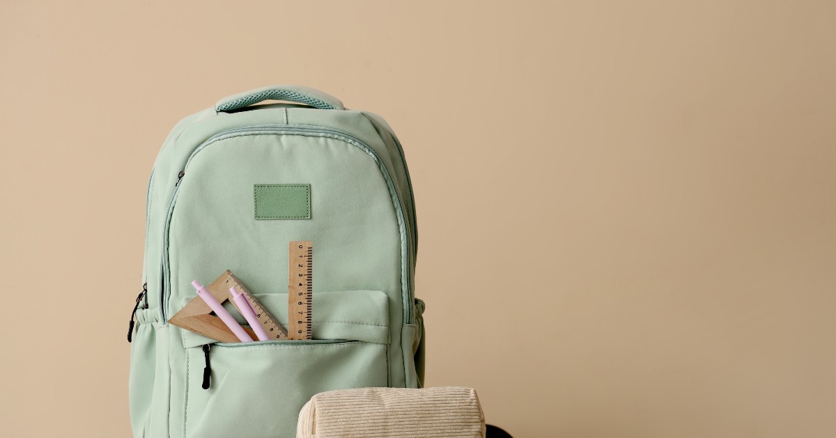 Un ghiozdan verde deschis stă pe o masă din lemn, având rigle și pixuri ieșite din buzunarul frontal. Lângă rucsac se află un penar bej textil și un calculator de birou verde, pe un fundal minimalist în tonuri calde.