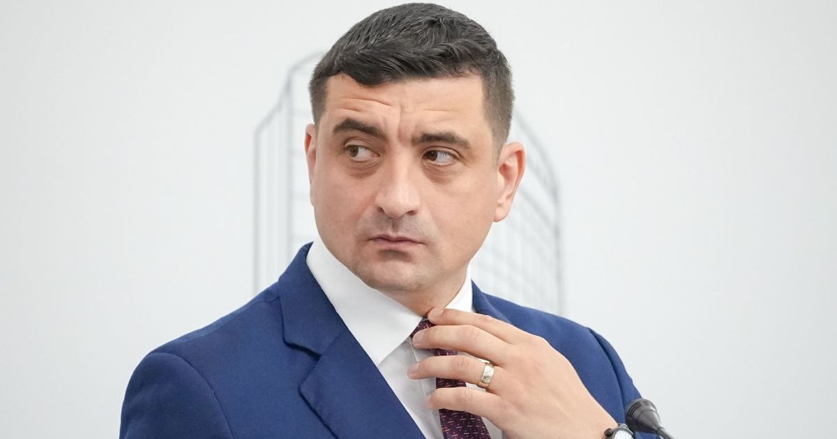 George Simion, îmbrăcat cu un costum albastru, în timp ce își aranjează cravata, pe fundalul unui perete alb