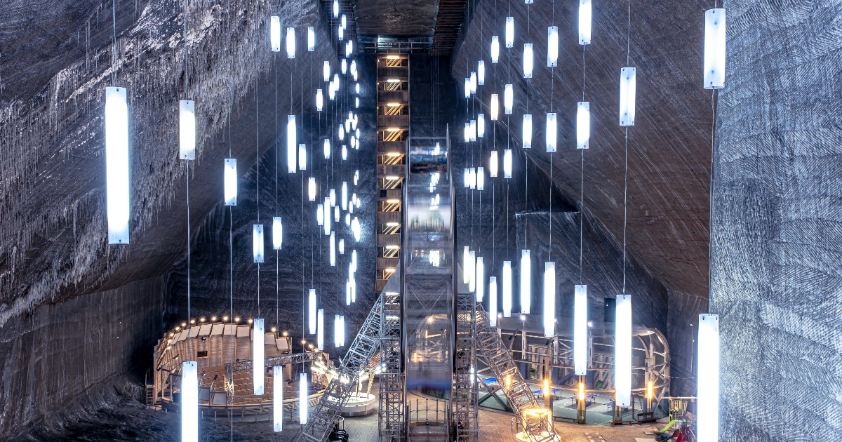 Imaginea suprinde interiorul spectaculos al Salinei Turda, cu pereții masivi din sare naturală și texturată. Spațiul larg, subteran, este iluminat modern de o multitudine de neoane verticale suspendate de tavan, care ghidează privirea către un lift central și zona de agrement de la baza camerei.