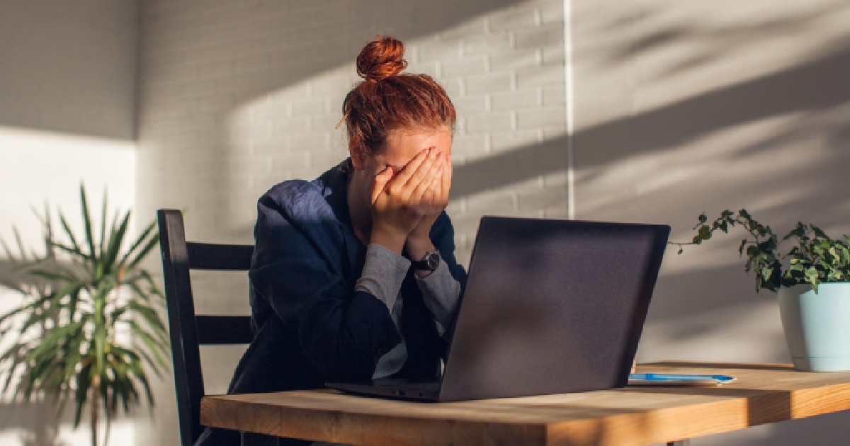 Imaginea surprinde o tânără cu părul roșcat strâns în coc, așezată la un birou în fața unui laptop, care își acoperă fața cu mâinile într-un gest de epuizare sau stres, într-o încăpere luminată natural de razele soarelui.