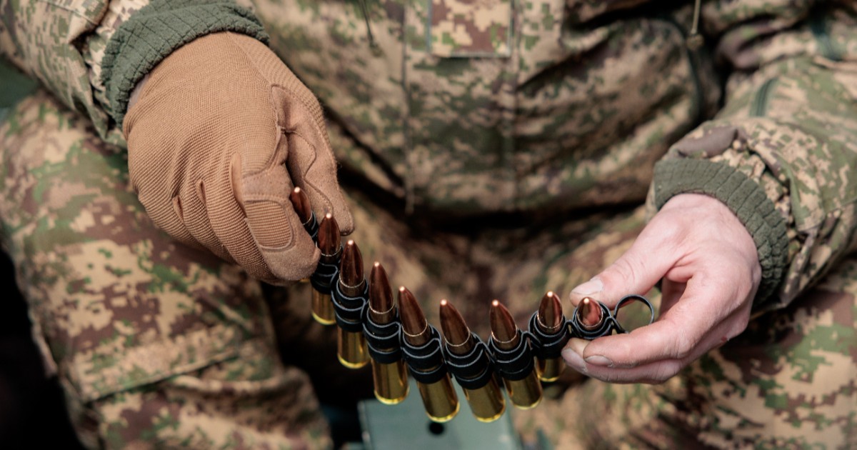 Un militar îmbrăcat în costum de camuflaj, în timp ce stă jos ți ține un cartuș de gloanțe în mână