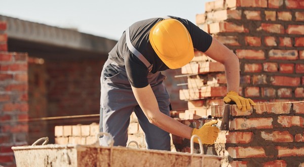Un muncitor în construcții care poartă cască galbenă de protecție și mănuși, lucrând la zidirea unui perete din cărămidă roșie pe un șantier.