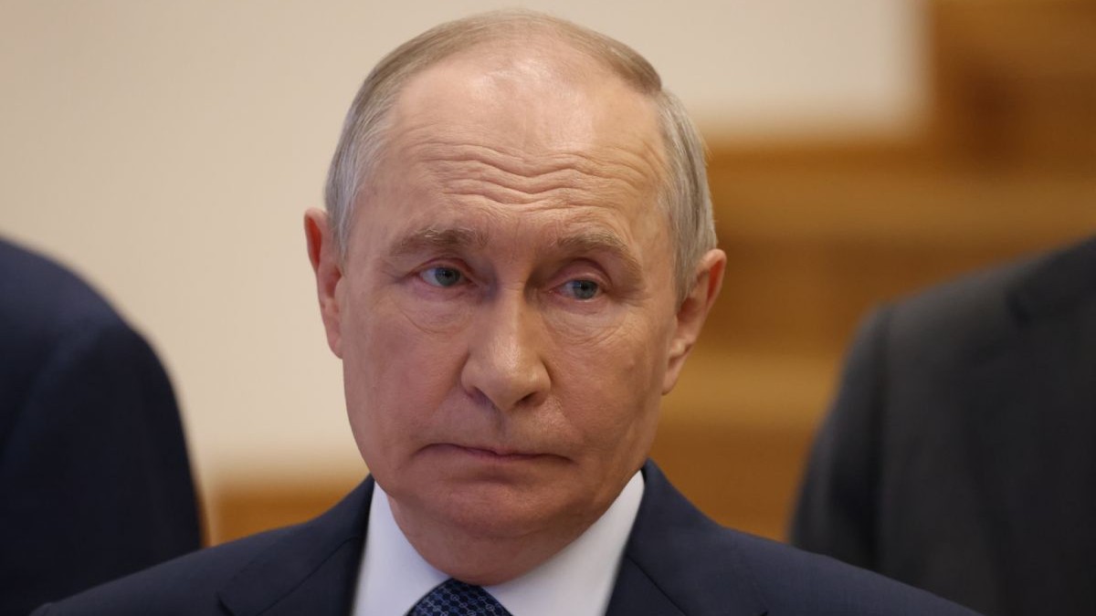 Vladimir Putin, îmbrăcat într-un costum negru, cămașă albă și cravată, cu fundal de culoare maro în blur