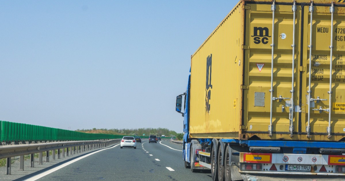 Imaginea surprinde un camion albastru ce transportă un container galben, rulând pe o autostradă alături de alte autoturisme, sub un cer senin.